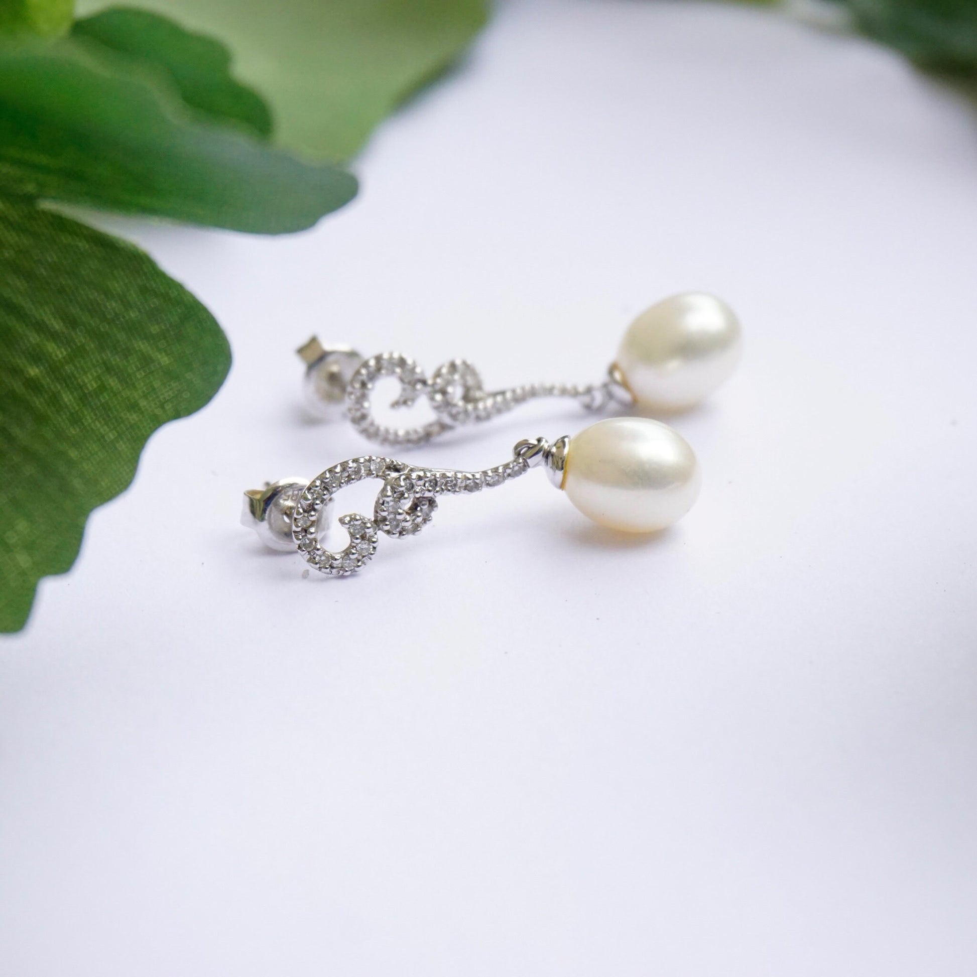 Vintage 14K white gold diamond and pearl drop earrings with delicate chain and stud design, set against green leaves on white background.