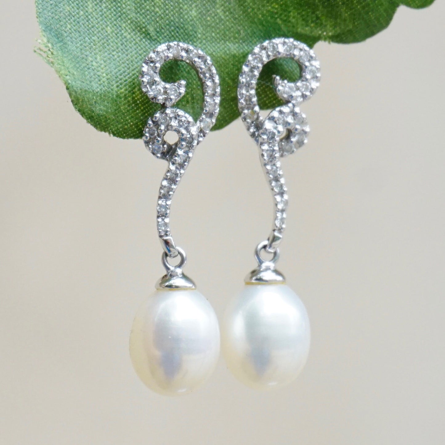 Vintage 14K white gold diamond and pearl drop earrings featuring delicate diamond swirl accents and lustrous oval pearl dangles, set on white gold stud posts against a soft green fabric background.