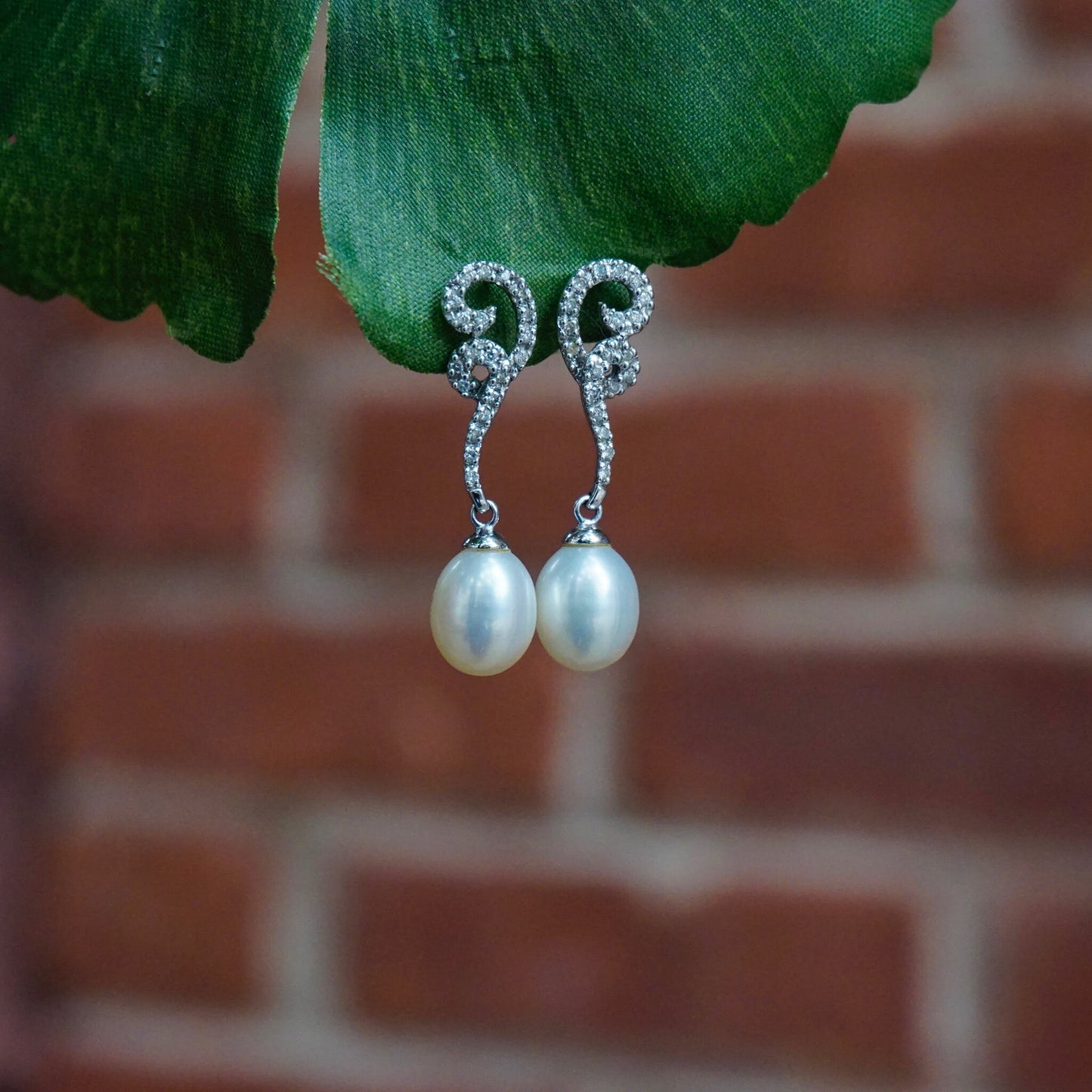 Vintage 14K white gold diamond and pearl drop earrings featuring delicate pearl drops suspended from sparkling diamond-accented swirl posts against a blurred brick background and lush green leaf.