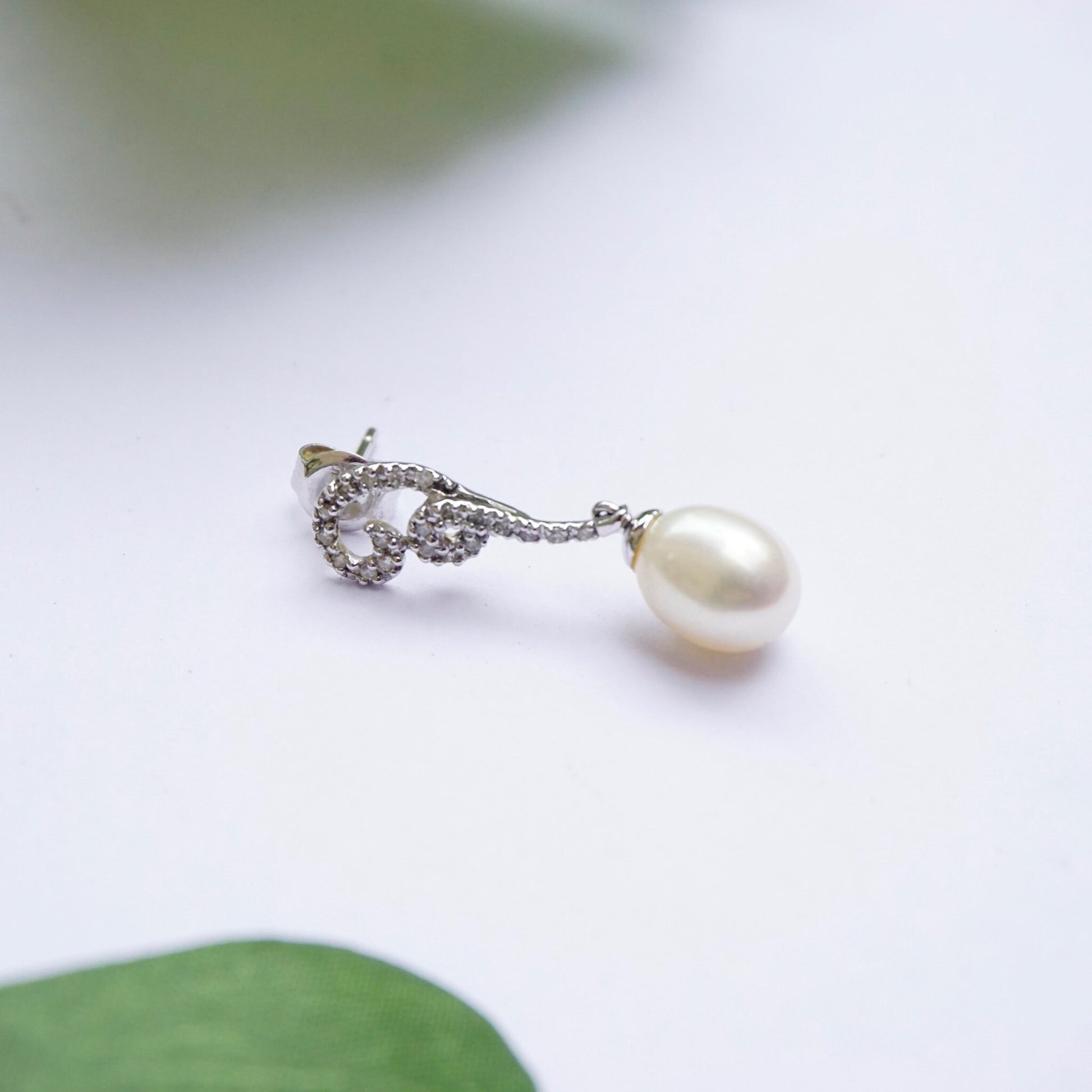 Vintage 14K white gold diamond and pearl drop earrings featuring delicate diamond accents on the stud and a lustrous pearl dangling elegantly below, photographed on a clean white background with a soft green leaf providing a subtle natural accent.