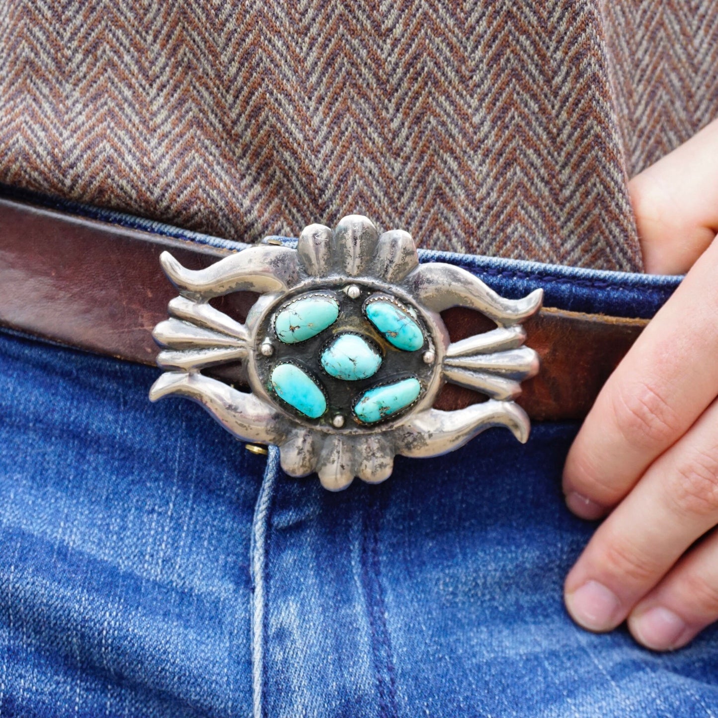 Vintage Navajo sterling silver and turquoise belt buckle featuring a central round turquoise cabochon surrounded by silver petals, set on a blue denim background held by a person's hand.