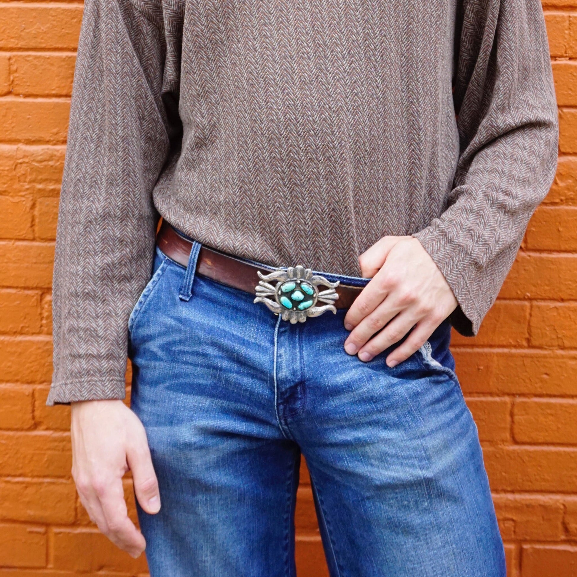 Vintage turquoise and sterling silver Native American belt buckle worn with jeans against a brick wall background.