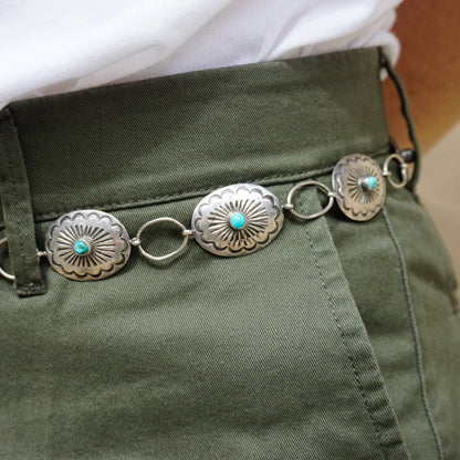 Vintage Native American sterling silver and turquoise concho belt with adjustable length and accent turquoise stones set in elaborately engraved silver conchos, displayed on green fabric, close-up view showing intricate silverwork details of old pawn style jewelry.