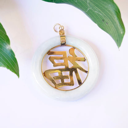 Vintage 14K gold jade Chinese symbol pendant featuring a large, light green jade disc with a cut-out Chinese character overlaid in 14K gold, hanging from a gold bail against a white background with green leaves.