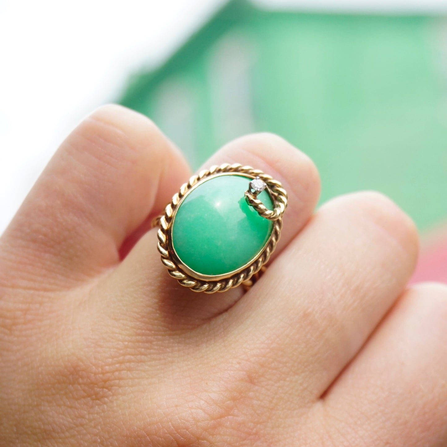 Vintage 14K gold jade and diamond cocktail ring featuring a large green jade stone accented by a single diamond, set in an intricate twisted gold band, shown on a hand against a green background.