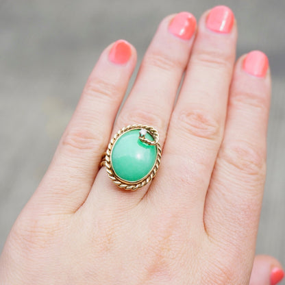 Vintage 14K gold cocktail ring with oval green jade cabochon center stone and twisted rope-style gold setting, accented by a single round brilliant cut diamond, shown on a hand with coral-painted fingernails against a beige background.