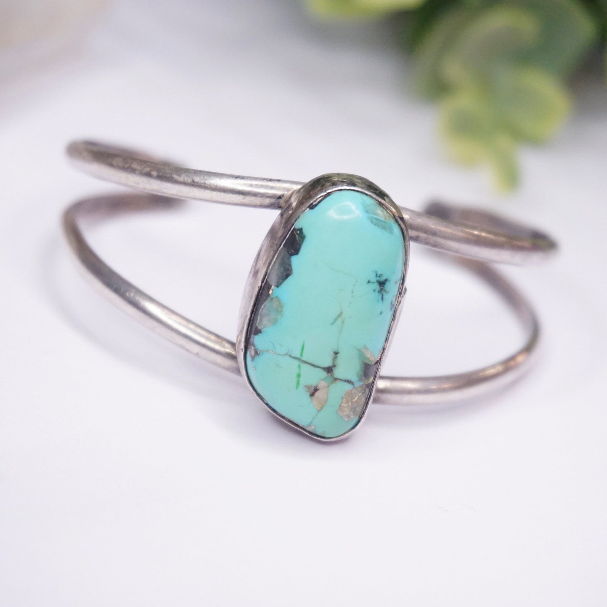 Vintage silver cuff bracelet featuring an oval turquoise stone, set on a light background with greenery.