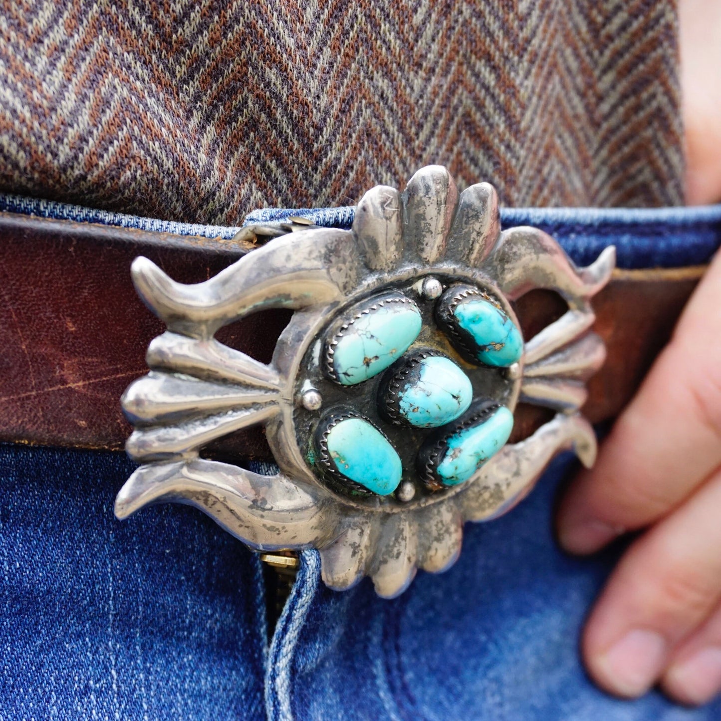 Vintage Navajo sterling silver turquoise belt buckle featuring multiple oval turquoise stones set in an ornate silver design resembling a sunburst or spider, held by a person wearing blue jeans. The belt buckle shows an antique, weathered patina on the silver.