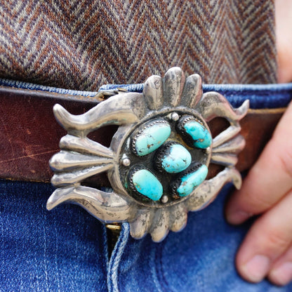 Vintage Navajo sterling silver turquoise belt buckle featuring multiple oval turquoise stones set in an ornate silver design resembling a sunburst or spider, held by a person wearing blue jeans. The belt buckle shows an antique, weathered patina on the silver.