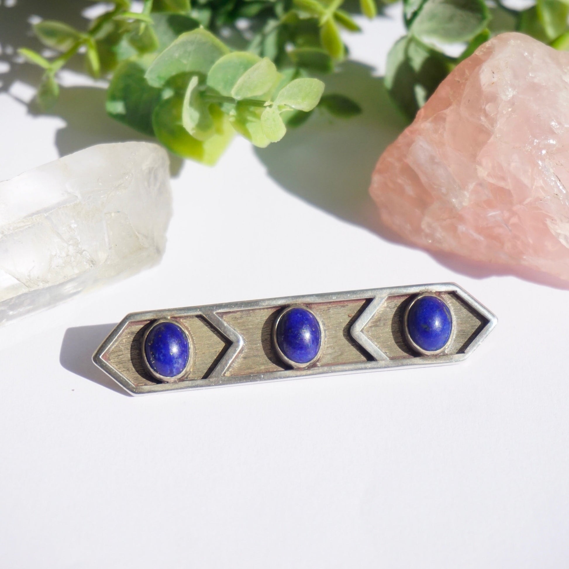 Vintage lapis lazuli and sterling silver brooch featuring three round lapis lazuli gemstones set in a minimalist rectangular silver design, displayed with rose quartz crystal and greenery in the background.