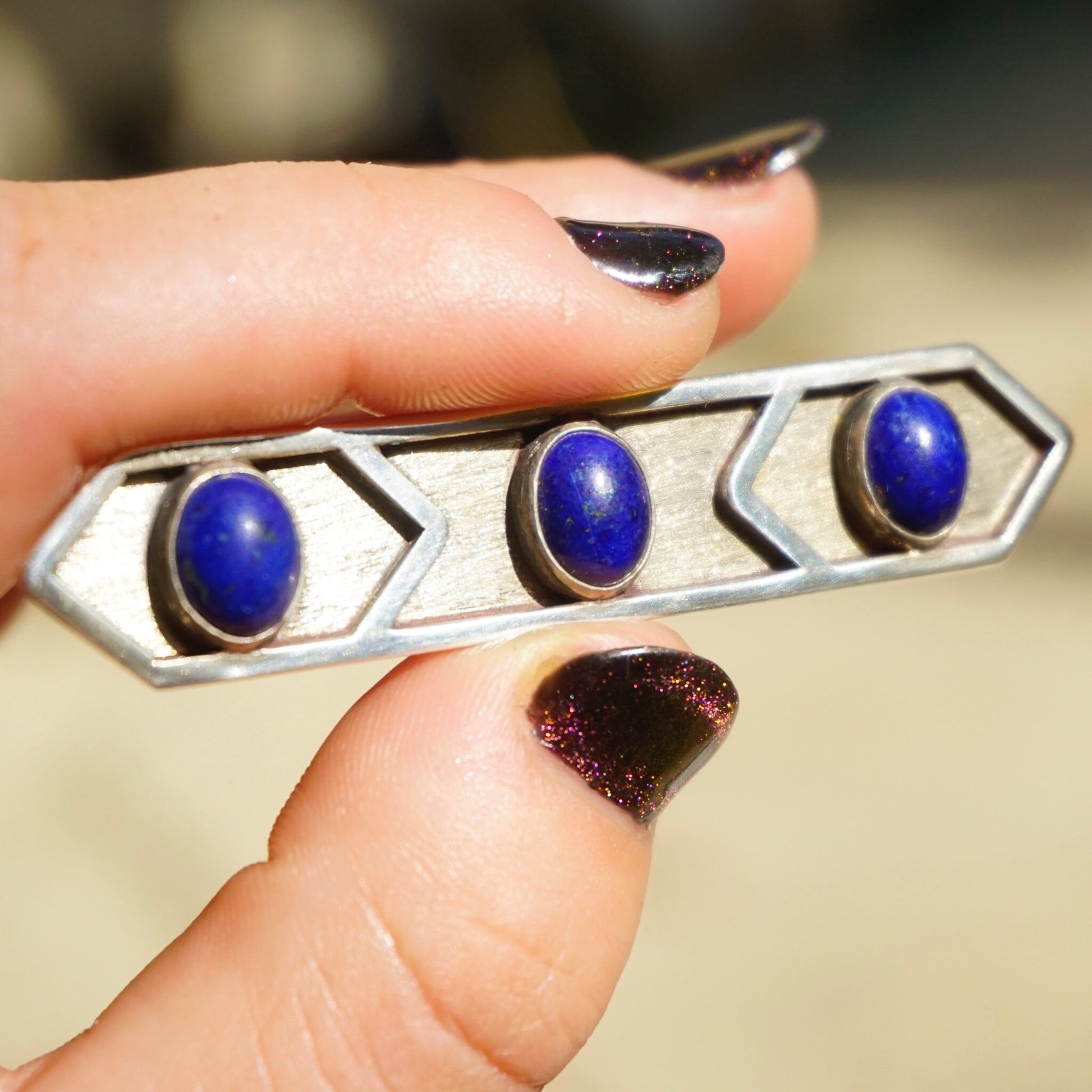 Vintage lapis lazuli and sterling silver three-stone brooch held in fingers with dark nail polish, featuring three round lapis stones set in minimalist hexagonal silver design.