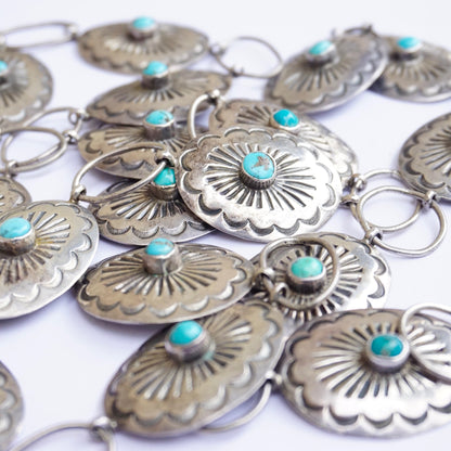 Vintage Native American sterling silver concho belt with turquoise accents. Adjustable silver belt features intricate floral designs on conchos inlaid with small turquoise stones. Old pawn jewelry crafted by skilled indigenous artisans. Close-up view of the detailed silver work and vibrant blue turquoise cabochons on a white background.