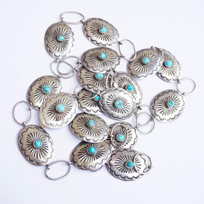 Vintage Native American sterling silver concho belt with turquoise accents, featuring multiple round conchos linked together, each decorated with an embossed flower design and a central turquoise stone, on an adjustable silver chain allowing the belt to be worn at various sizes, a beautiful example of traditional Old Pawn Native American jewelry craftsmanship.