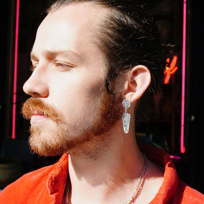 The image shows a close-up portrait of a man with a reddish beard, mustache and sideburns. He has his eyes closed and appears to be wearing a red shirt or jacket. The man has silver dangle earrings with a unique swirl design hanging from his ears. The background is dark, putting the focus on the man's face and jewelry.