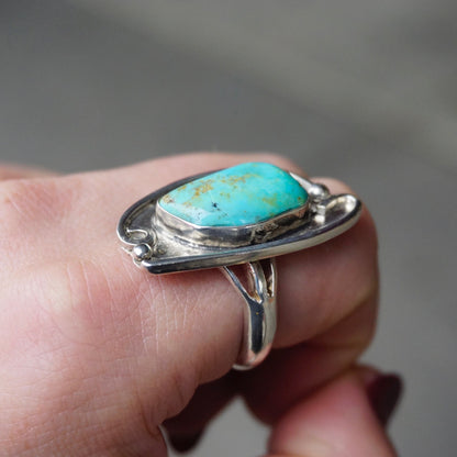 Vintage turquoise sterling silver ring featuring a large natural turquoise stone in an oxidized silver setting, reminiscent of Native American jewelry designs, shown worn on a finger against a gray background.