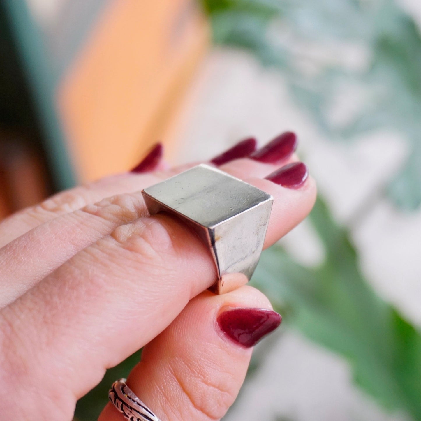 Vintage sterling silver signet ring with thick rectangular face held between fingers with red nail polish, against blurred green leaf background.