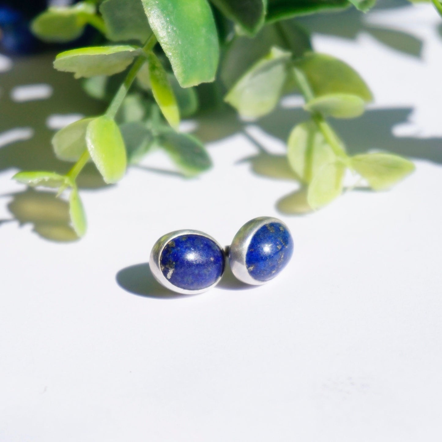 Vintage lapis lazuli stud earrings in sterling silver, resting on a white surface with green leaves in the background. The round, deep blue lapis lazuli gemstones are bezel-set in minimalist 925 silver stud earring settings, creating an elegant accent jewelry piece.