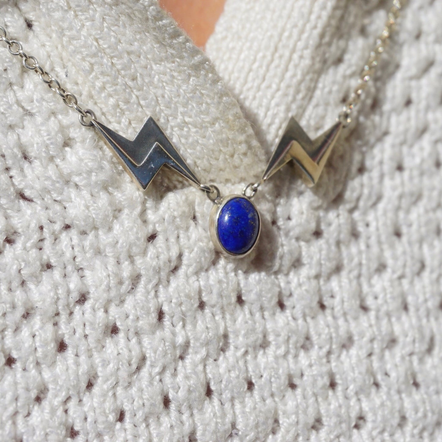 Vintage lapis lazuli silver necklace featuring a unique lapis stone pendant in an oval shape, accented by two silver lightning bolt charms on a delicate silver chain, showcased on a cozy beige knit sweater background.