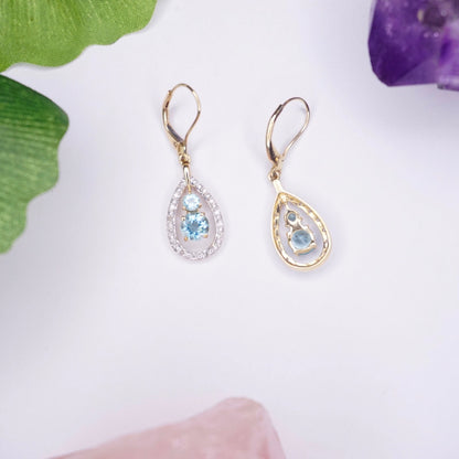 14K gold teardrop earrings with aquamarine center stones surrounded by accent diamonds, shown on white background with green leaves and purple crystals.