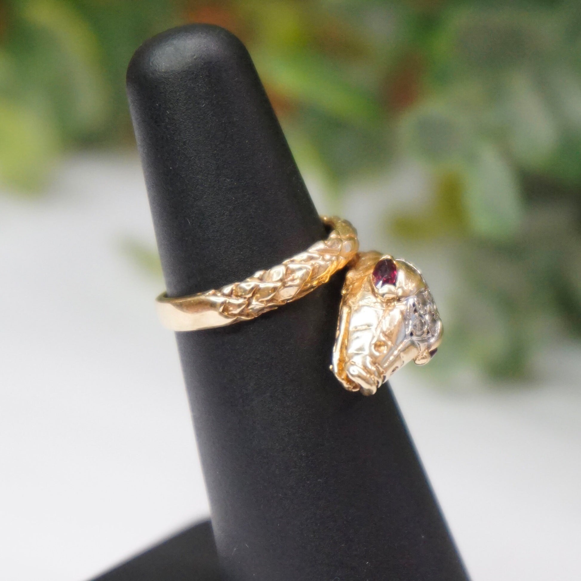 Vintage 14K yellow gold snake ring with diamond-encrusted body wrapping around the finger, accented by two ruby eyes, displayed on a black ring stand with blurred green foliage background.