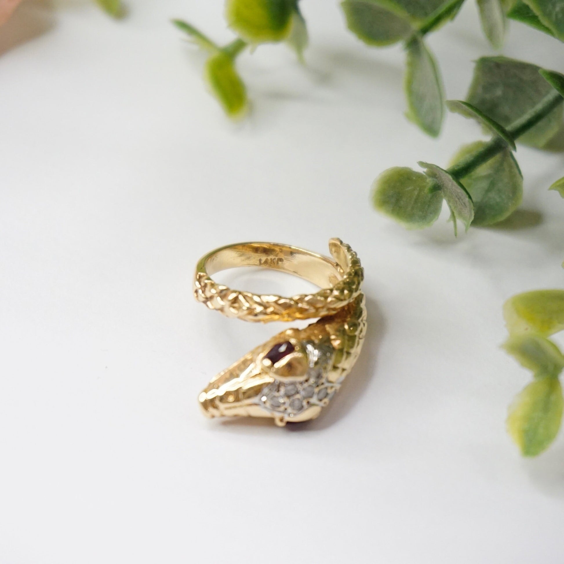 Vintage 14K gold wrap around snake ring with diamond-encrusted body and ruby eyes, set against a background of green leaves.