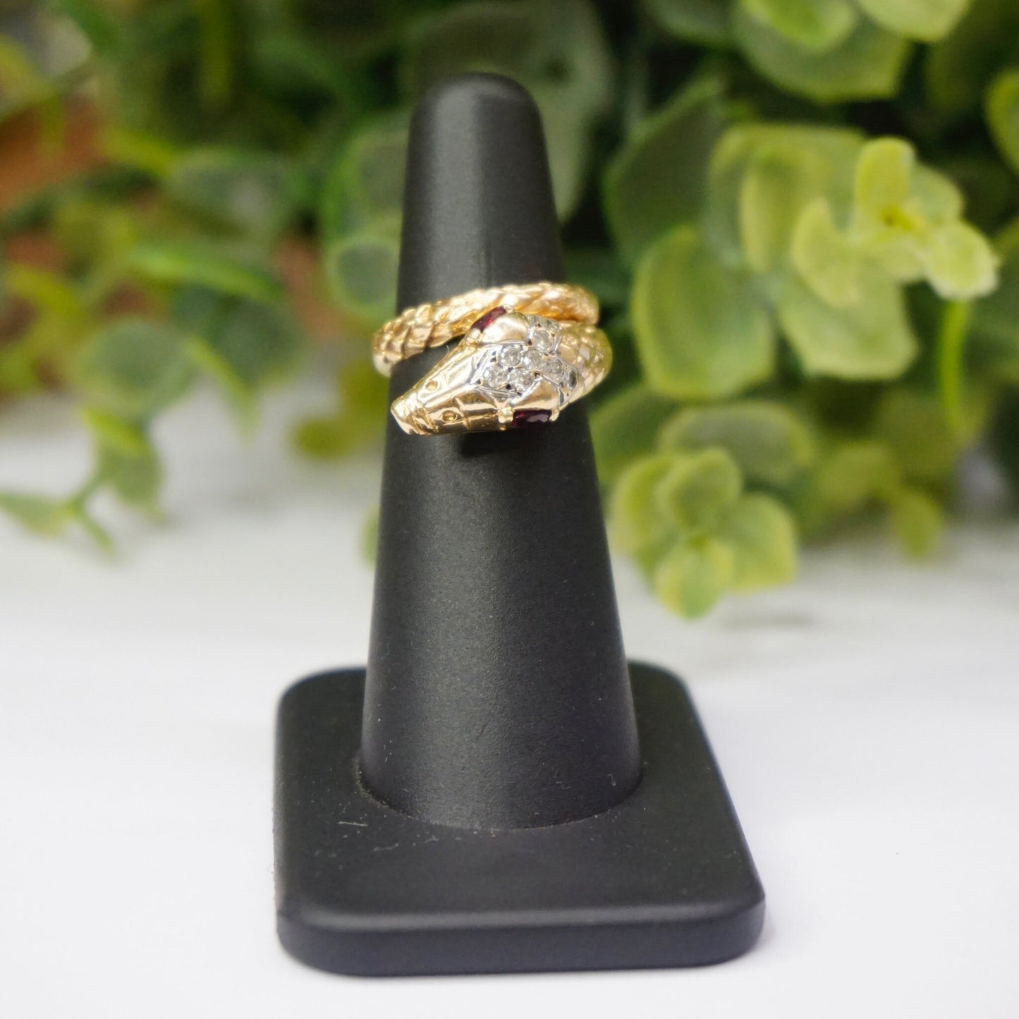 Vintage 14K gold snake ring featuring ruby eyes and diamond accents, wrapped around a black jewelry display stand, shown with greenery in the background.