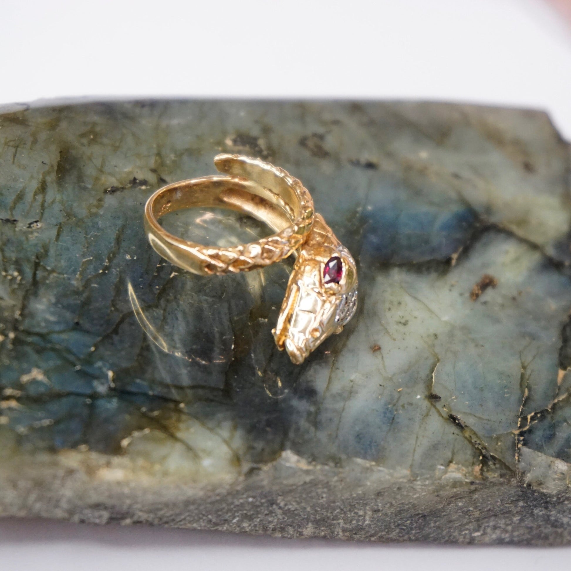 Vintage 14K gold wrap around snake ring featuring ruby eyes and diamond encrusted body, displayed on a textured gray stone surface.