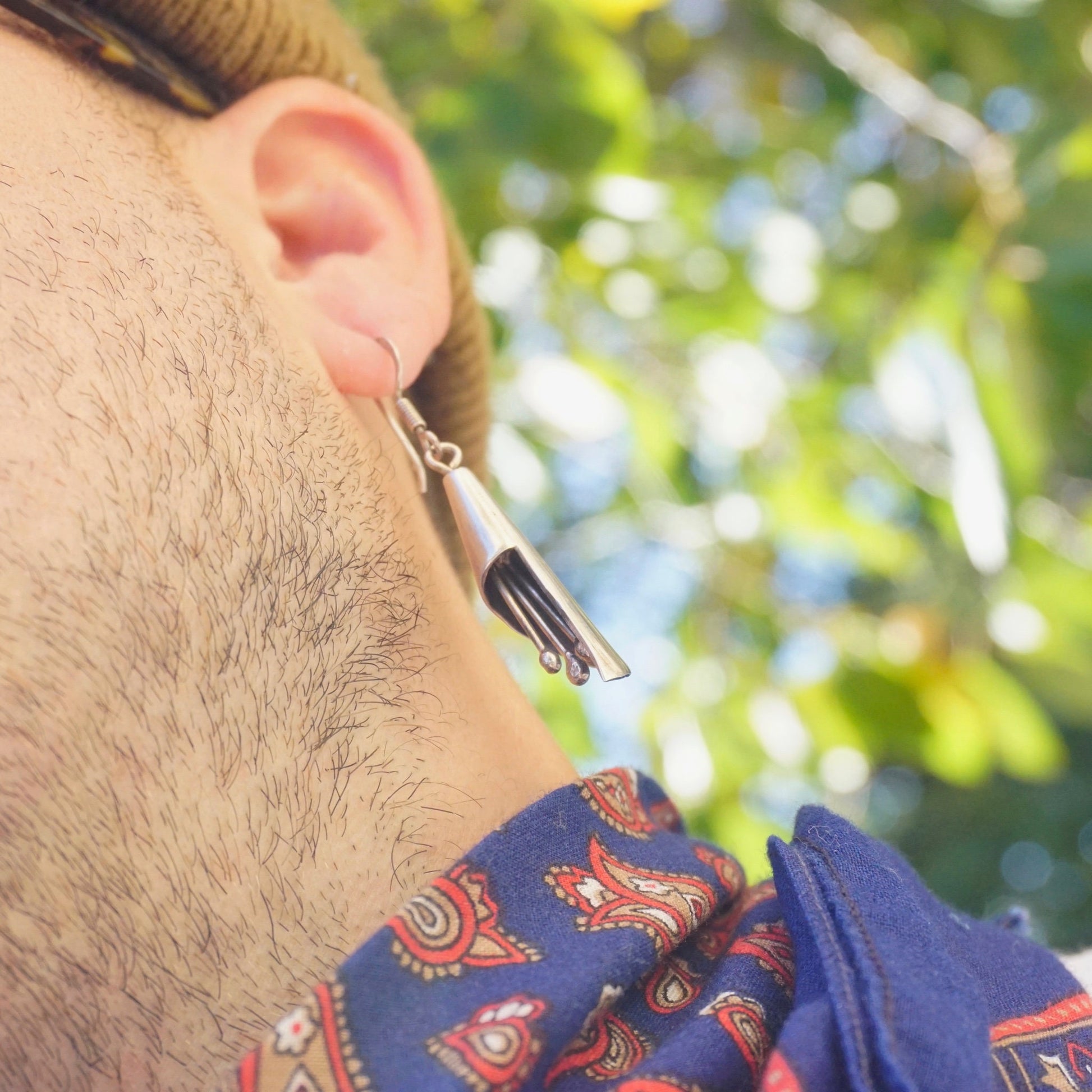 The image shows a close-up of a man's ear and neck, with silver dangle earrings hanging from the ear. The earrings have a modernist design with multiple dangling elements that would jingle or chime when moved. The background is blurred green foliage, suggesting an outdoor setting on a sunny day.