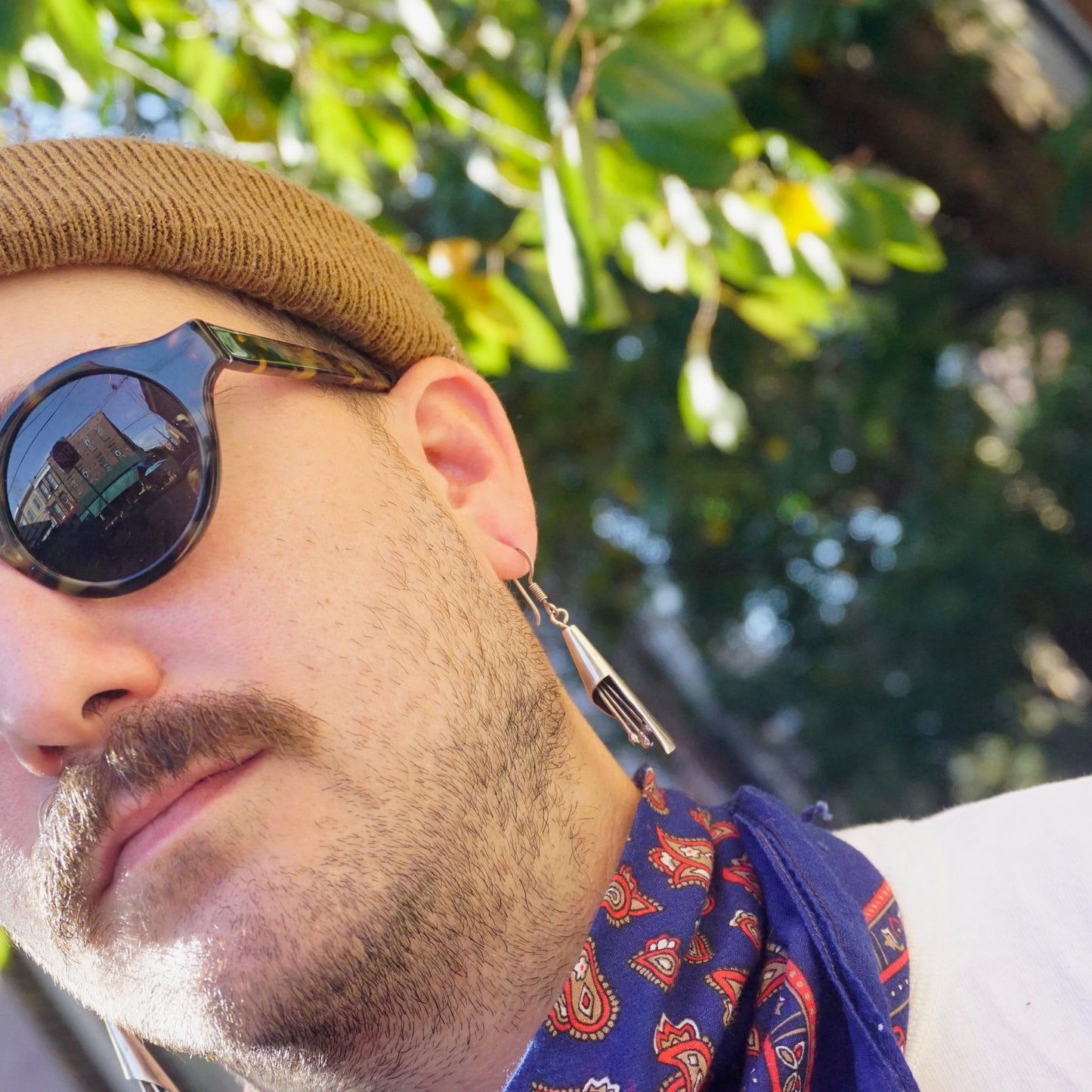 Close-up portrait of a man wearing sunglasses and a hat with a leaf pattern, looking to the side in an outdoor setting with trees in the background.