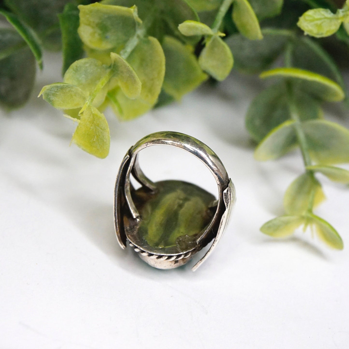 Vintage turquoise sterling silver ring with natural stone and intricate wing design, set against a backdrop of green leaves. Native American inspired 925 silver jewelry.