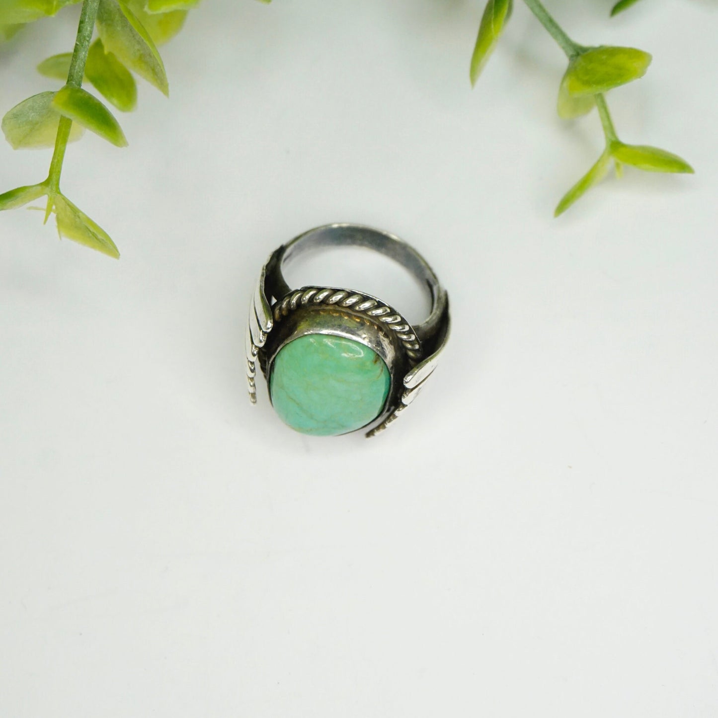 Vintage turquoise and sterling silver ring with rope design band and round green turquoise stone set against white background with green leaves