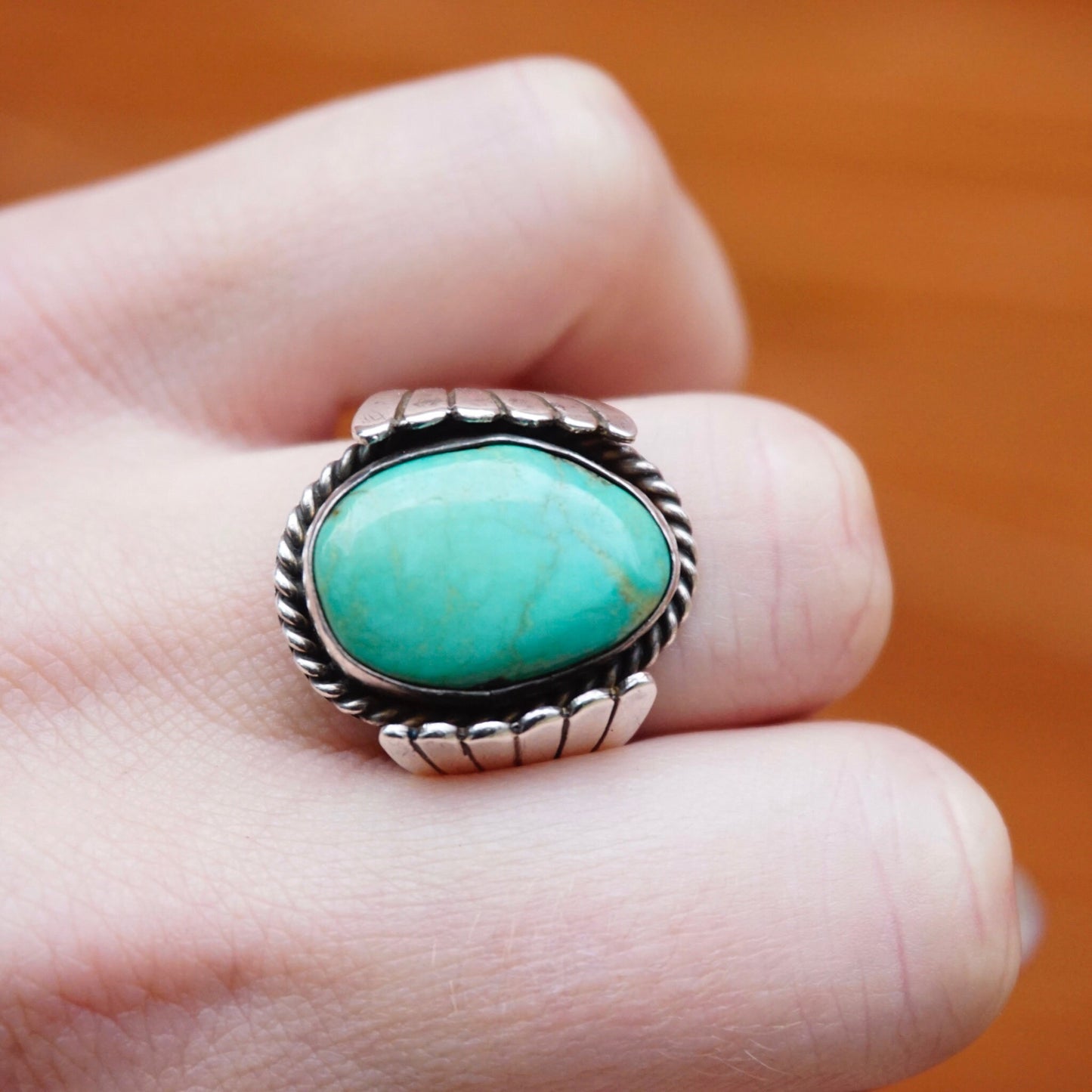 Vintage turquoise and sterling silver ring featuring a natural turquoise stone set in an ornate silver bezel with a twisted rope design along the band, held in a person's fingers against an orange background.