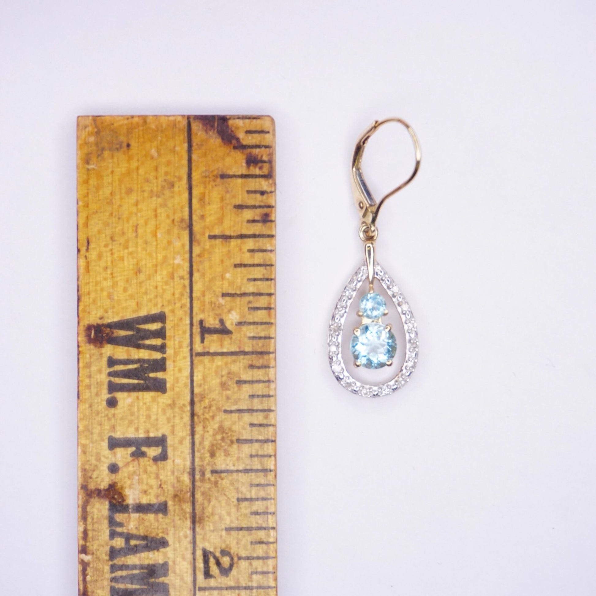 14K gold teardrop earrings featuring an aquamarine gemstone surrounded by diamond accents, shown next to a wooden ruler for scale.