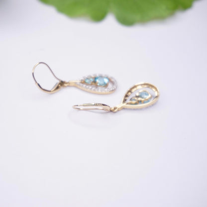 Vintage 14K gold tear drop earrings with aquamarine and accent diamonds, dangle style with light blue gemstone centers surrounded by small diamonds, hanging on gold lever back earring hooks, photographed on white background with blurred green leaf in the background.