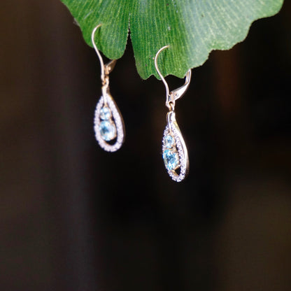 Vintage 14K gold tear drop earrings with aquamarine center stones and diamond accents hanging from green leaf, closeup view on black background