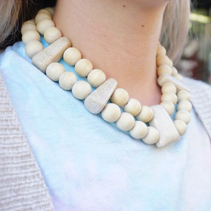 Vintage Gerda Lynggard Monies chunky wooden beaded necklace with two strands featuring large round wooden and stone beads in neutral tones worn around a woman's neck, closeup view of the mid-century statement collar necklace against a light blue sweater.