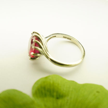 Vintage 10K white gold ruby cocktail ring with deco spiral setting and raised bright red stone on a green leafy background.