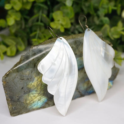 Vintage mother of pearl angel wing earrings with iridescent shell hanging from silver hooks against green foliage background