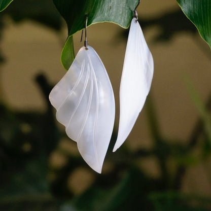 White mother of pearl angel wing earrings hanging from green leaves, creating an ethereal and delicate vintage jewelry look with iridescent shell material and silver dangle design.