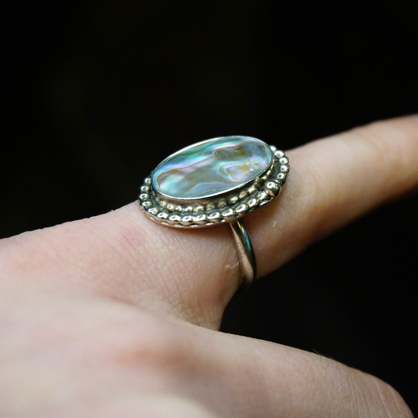 Vintage sterling silver abalone ring with textured band and petite oval abalone shell cabochon on a finger, against a black background.