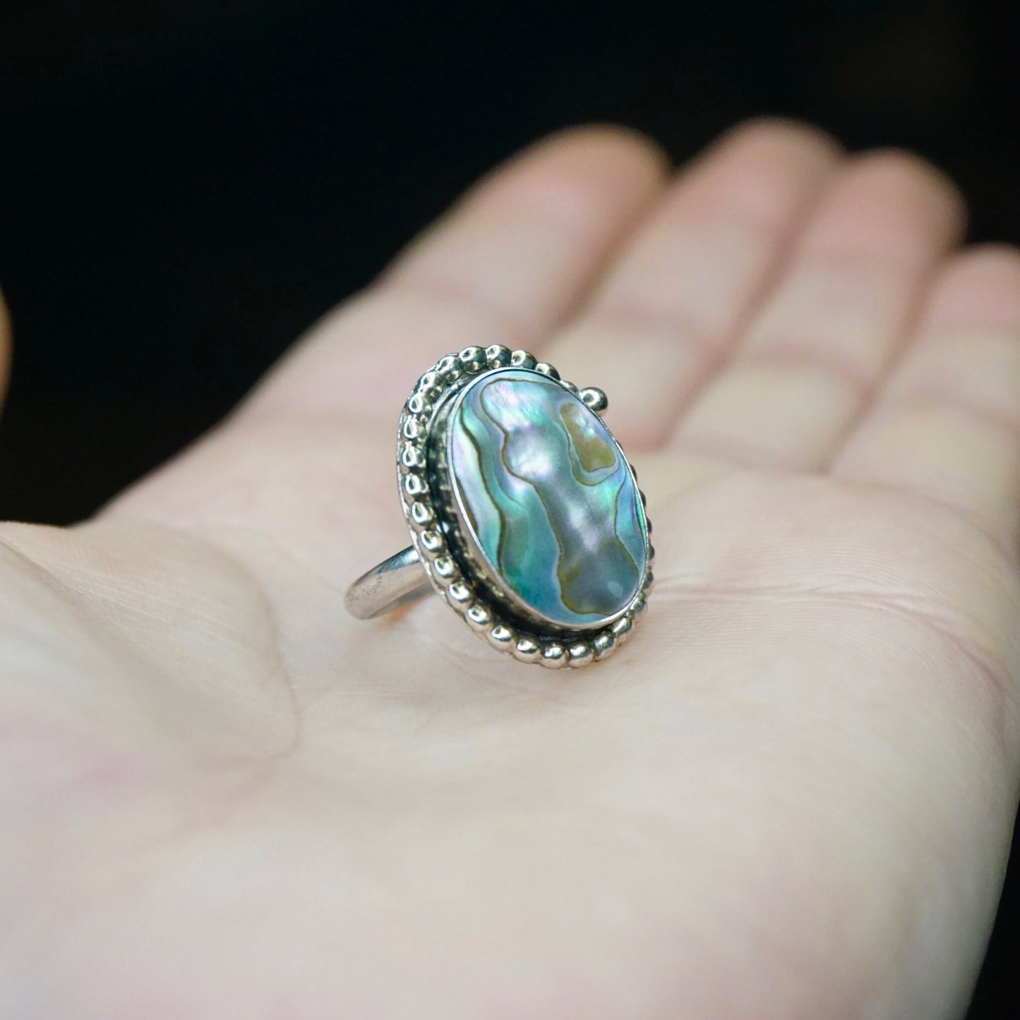 Vintage sterling silver ring with oval abalone shell cabochon set in textured bezel on an open hand, displaying the iridescent blue-green colors of the abalone against the oxidized silver setting.