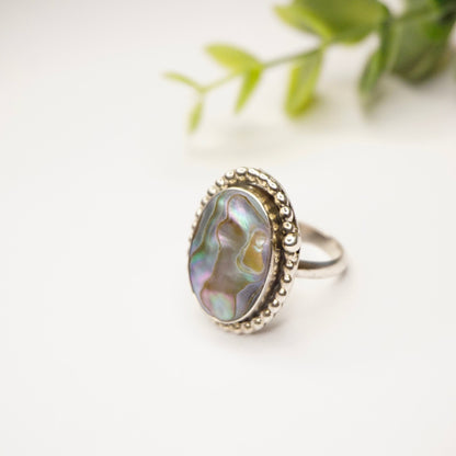 Vintage sterling silver abalone ring with textured band and iridescent oval abalone inlay, displayed on white background with greenery accent.