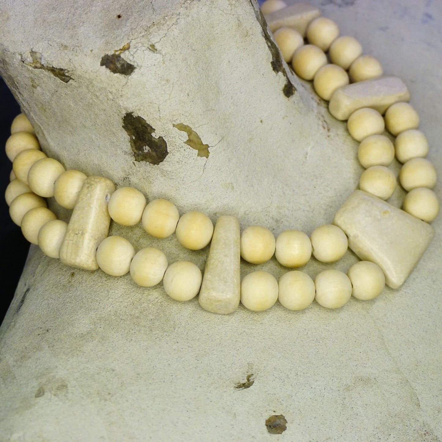 Vintage wooden bead necklace with two strands of chunky, round beads arranged in a collar style, contrasted against a textured, light-colored stone surface with small specks of debris.