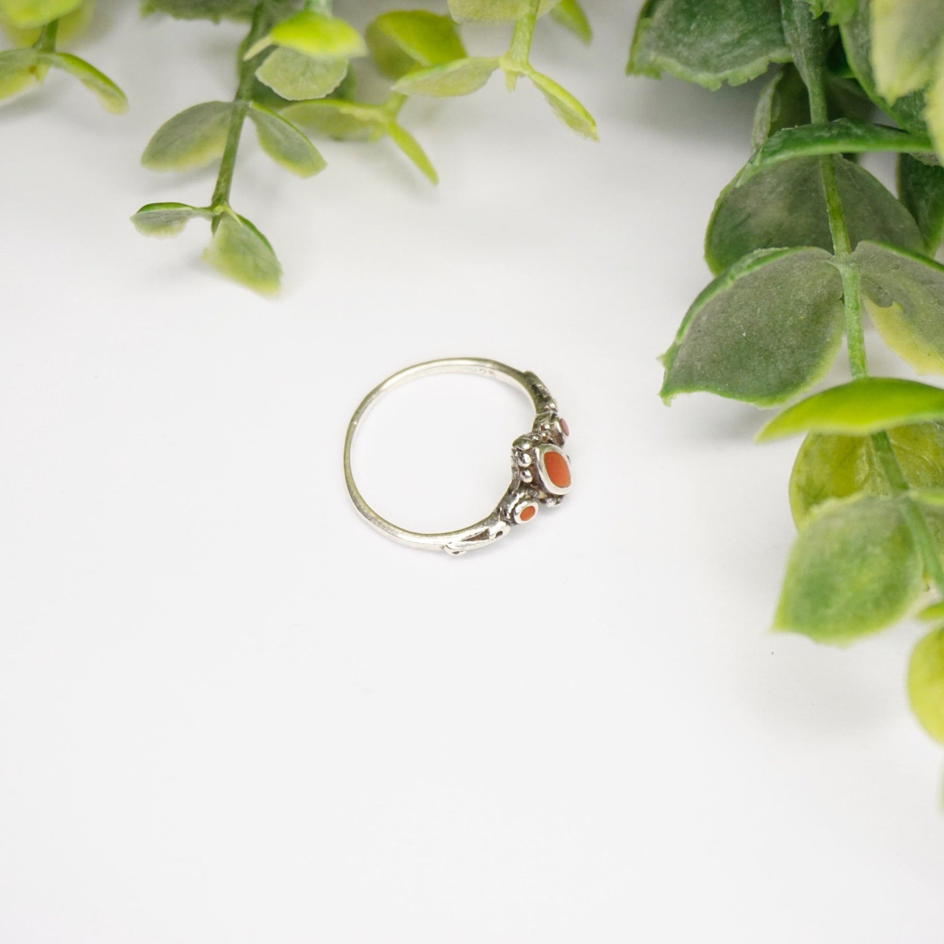 Vintage sterling silver ring with red stone or glass accent, featuring intricate swirl details on a thin band, shown on a white background with green foliage.