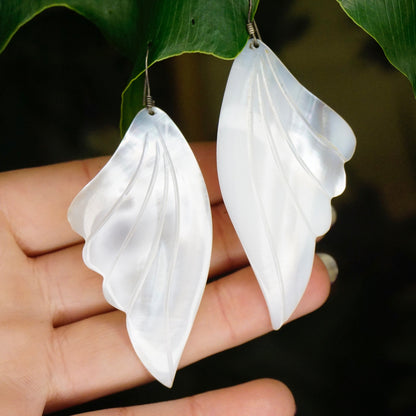 Vintage mother of pearl angel wing dangle earrings, iridescent shell carved into feather shapes, set in 925 silver, held in open palm against green leaf background.