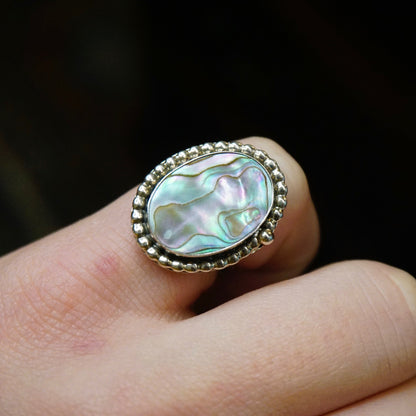 Vintage sterling silver ring with an oval-shaped abalone shell inlay surrounded by a textured beaded border, shown on a person's finger against a black background.