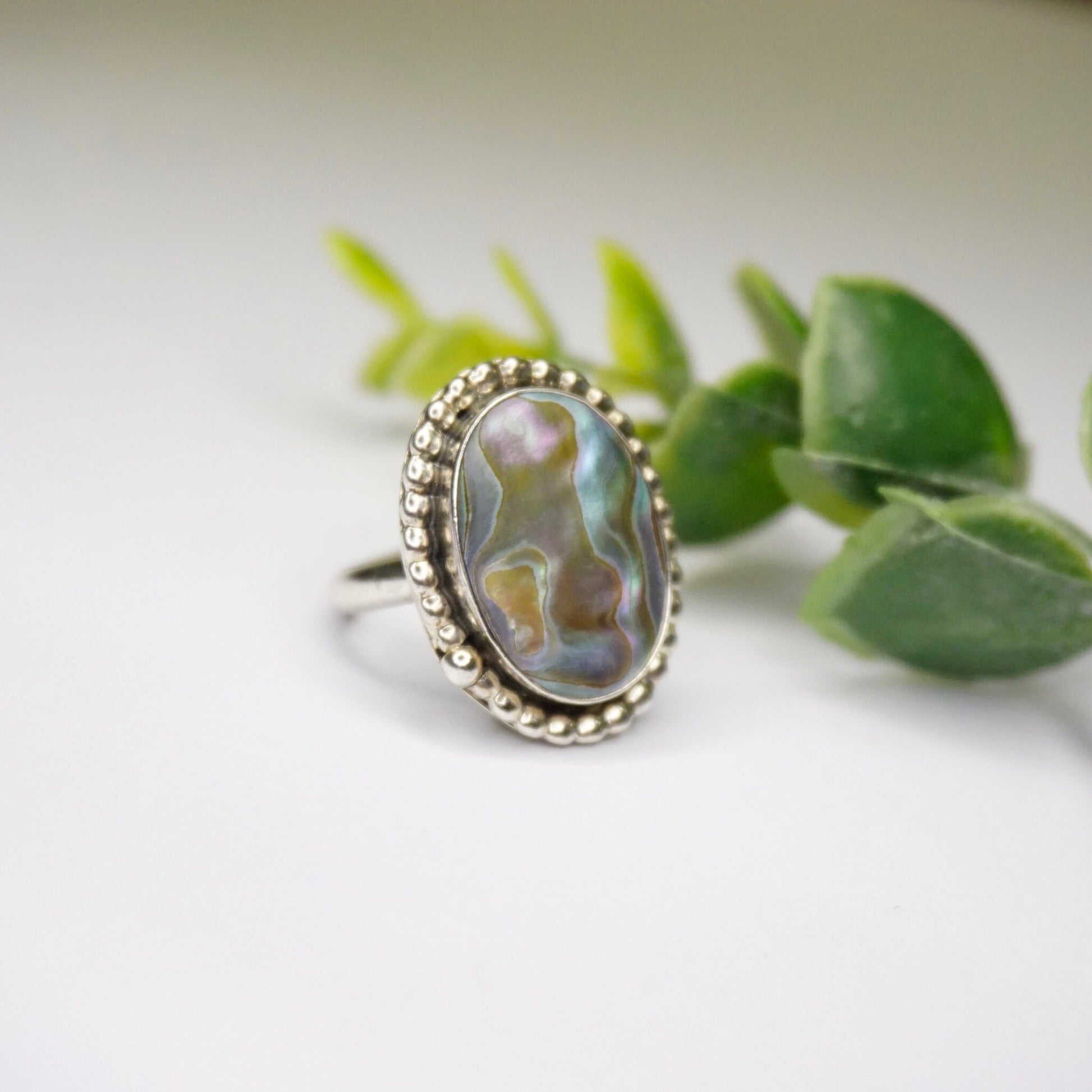 Vintage sterling silver abalone ring with textured design and oval-shaped abalone shell, displayed on white surface with green leaves in background