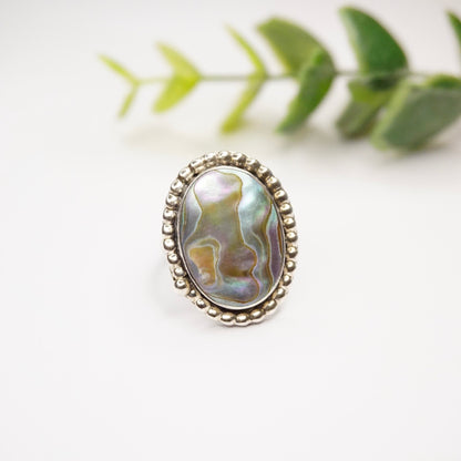 Vintage sterling silver abalone ring with textured band and petite oval-shaped iridescent abalone stone, displayed on white background with green leaves.