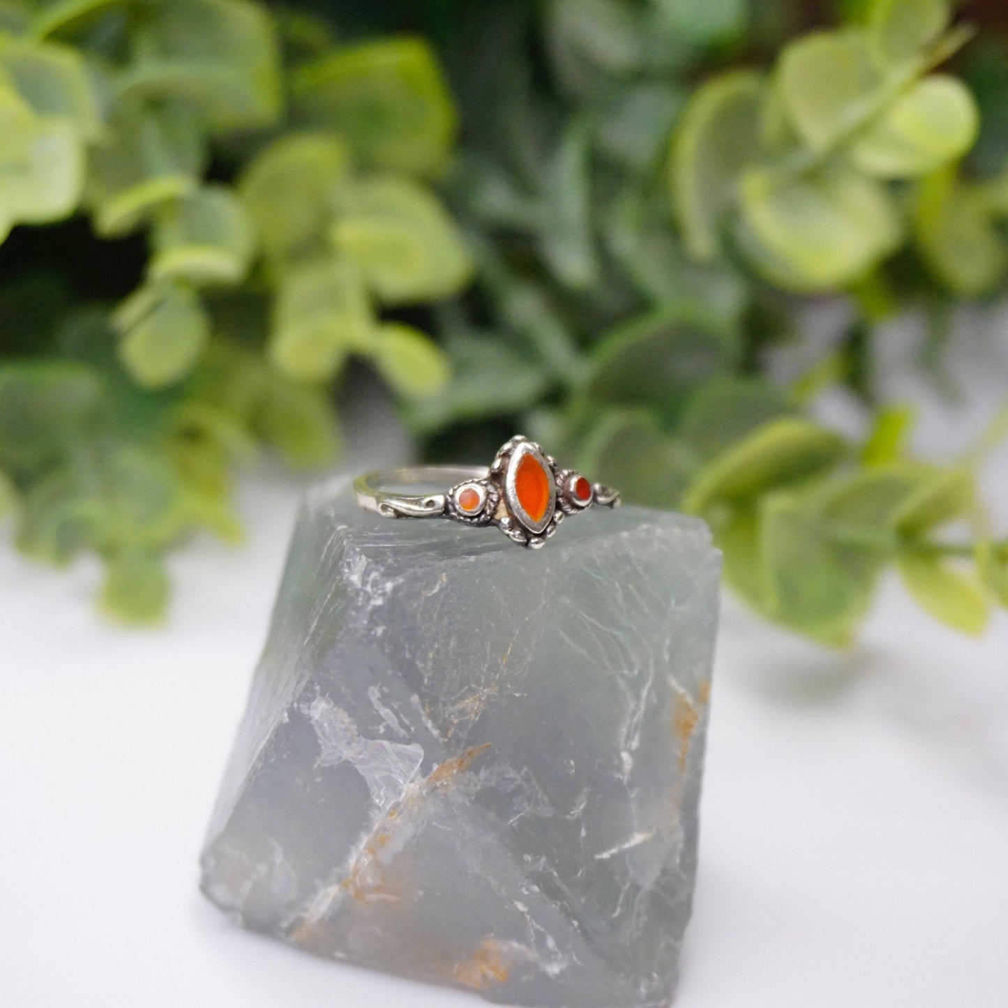 Vintage sterling silver ring with red stone or glass accent, featuring delicate swirl details on a thin band, placed on gray stone with green foliage in the background.