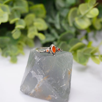 Vintage sterling silver ring with red stone or glass accent, featuring delicate swirl details on a thin band, placed on gray stone with green foliage in the background.